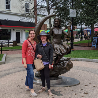Two women posing in front of a bronze statue of the Bewitched Statue in Salem, Mass, with a building and trees in the background.
