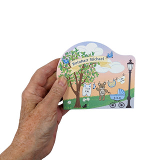 A hand holding a small, die-cut wooden keepsake box with a colorful cartoon illustration of a park scene and the names 'Kenneth Michael' printed on the front, set against a plain gray background.