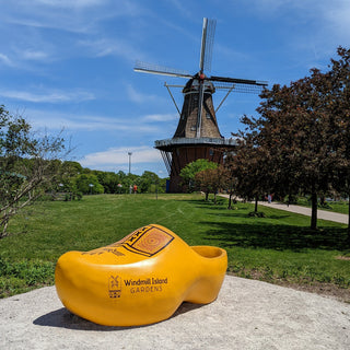 A large yellow clog-shaped bench sits on a concrete surface in a park, with a traditional Dutch windmill, green grass, and a blue sky with clouds in the background.