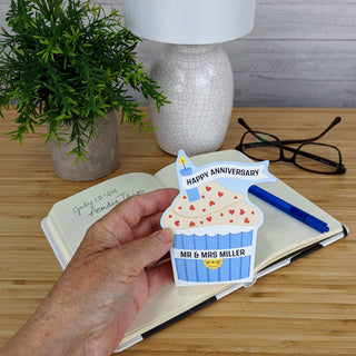 Hand holding a personalized anniversary card with a cupcake design on a wooden desk.