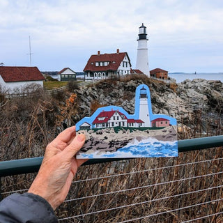 A hand holds wooden replica lighthouse and coastal house scene in front of a real outdoor coastal landscape with a lighthouse, rocks, ocean, and cloudy sky.