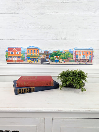 Colorful 3/4" thick wooden children's decor with a town illustration on a white surface with books and a plant.