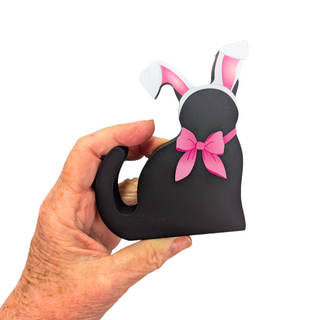 hand holding a wooden replica black cat that has a pink bow and bunny ears on a white background 