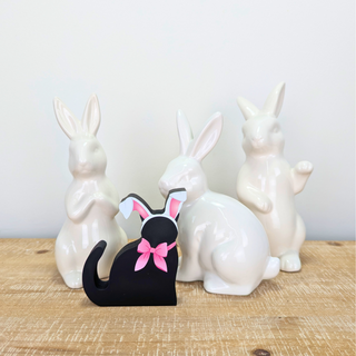 hand holding a wooden replica black cat that has a pink bow and bunny ears on a white background with white ceramic rabbits behind it 