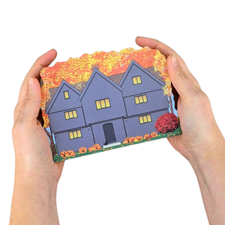 Hands holding a small 3/4" thick wooden replica of the Salem Witch House with autumn scenery