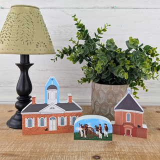A set of small wooden models of historic churches and a carriage scene, displayed on a wooden surface alongside a black table lamp and a potted green plant.
