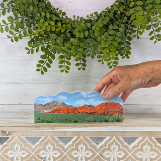 Hand holding a small landscape card with mountains and greenery against a white wall with green leaves.