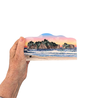 Hand holding a 3/4" wooden cut out replica of an illustration of a  with a scenic view of a beach and cliffs.