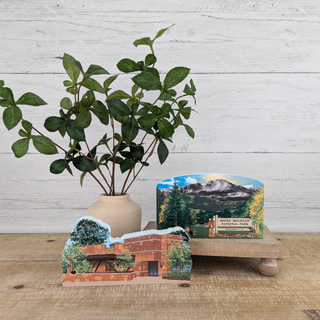 Stack of scenic paper models on a wooden surface with a plant in the background