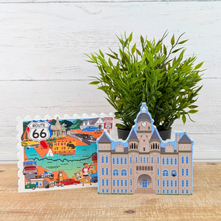 A colorful Route 66 map-shaped card and a 3D paper model of a historic building, styled on a wooden surface with a green potted plant against a white wood plank background.