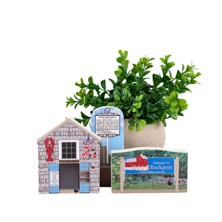 A decorative miniature village scene featuring a small shingled house, a blue signpost, a potted green plant, and a 'Welcome to Rockport' sign on a white background.