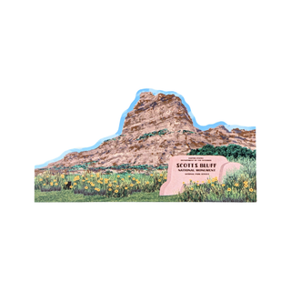 3/4" thick wooden replica with an illustration of a Scotts Bluff National Monument, featuring a large layered sandstone cliff, green grass, yellow wildflowers, and a pink monument sign on a white background.