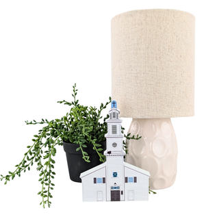 A decorative tabletop scene with a small white church figurine in front of a cream textured lamp base and beige drum shade, alongside a green trailing plant in a black pot, all on a white background.