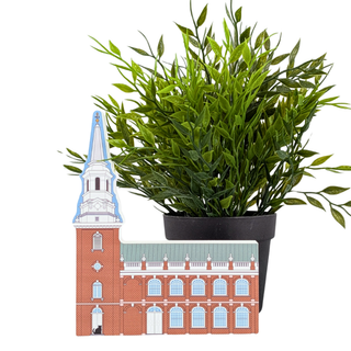 An artificial green leafy plant in a black plastic pot, positioned behind a decorative cutout of a red brick church with a white steeple and blue trim, set against a plain white background.