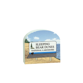 A decorative sign shaped like a 'Sleeping Bear Dunes National Lakeshore' entrance sign, featuring a beach, blue sky, lake, and a small black bear silhouette on a white signboard.
