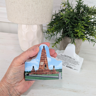 Hand holding a small 3/4" thick wooden  replica with a picture of a tower, with a lamp and plant in the background.