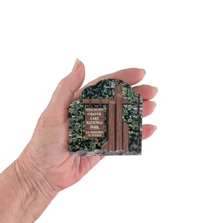 Hand holding a small 3/4" wooden replica with an illustration of the welcome to crater lake national park 