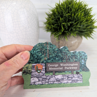Hand holding a 3/4" thick wooden illustration of a memorial Parkway sign with stone wall and trees on a white background