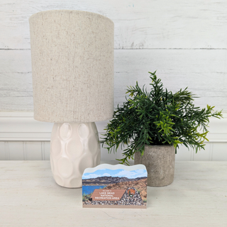 Lamp with beige shade, plant, and postcard on a light surface with white wooden background