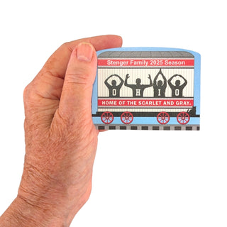 Hand holding a small wooden replica train car with 'Stenger Family 2025 Season' and 'Home of the Scarlet and Gray' text.