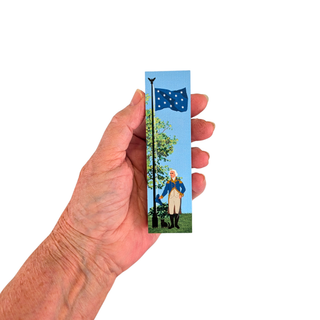 A hand holding a s3/4" thick wooden replica with an illustration of a blue flag, a tree, and a historical figure against a light blue sky, shown against a plain white background.