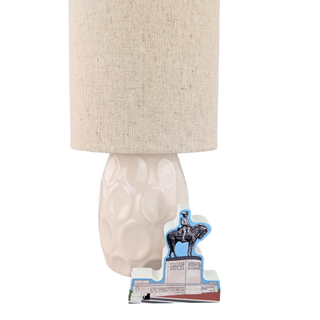A table lamp with a textured beige linen drum shade and a glossy white ceramic base featuring a raised bubble pattern. In front of the base sits a small die-cut decorative card with a cowboy on a horse illustration and printed text.