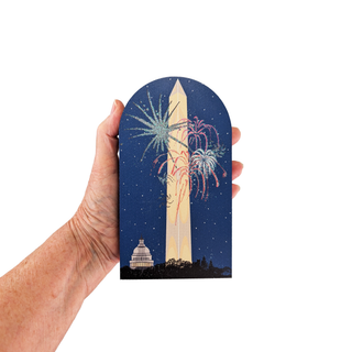 Hand holding a 3/4" thick wooden illustration of the Washington Monument and fireworks against a night sky.