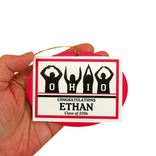Graduation-themed ornament with 'OHIO' and 'Congratulations Ethan Class of 2016' text, held by a hand against a textured beige background.