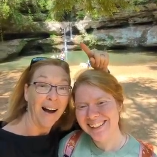 Two people posing for a selfie in front of a natural water feature with trees and rocks.
