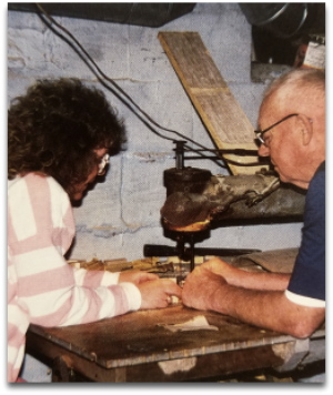 Two people working on a wooden object in a workshop setting.