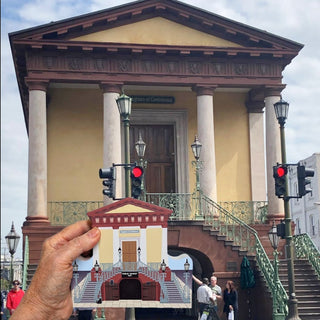 A hand holds a small printed wooden replica depicting the front facade of a classical building with columns, stairs, and red traffic lights in the foreground, set against a real outdoor scene of a similar building.