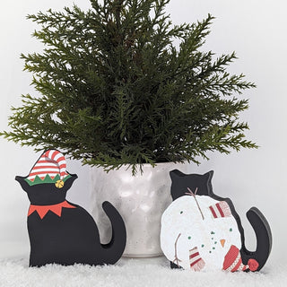 potted plant with two black cat wooden replicas, one with an elf hat and the other as a snowman 