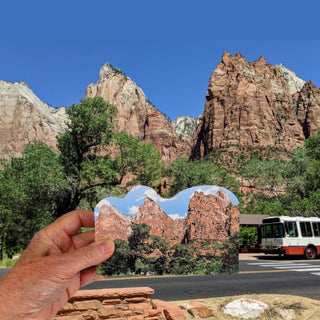 A hand holds a wooden replica  of Zion National Park under a blue sky, superimposed over a real outdoor scene with trees, a road, and a bus. The product appears to be the printed scenic sticker/print rather than the environment itself.