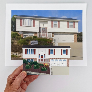 A hand holds a small printed illustration or mockup in front of a larger original photo of a house, both on a white background. The smaller printed piece shows a front view of the house with landscaping and a garage, while the larger photo shows the real house exterior under a blue sky.