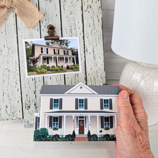 A hand holds a small printed house-shaped card or plaque in front of a rustic white wood panel background, with a larger photo of a house clipped above it and a white lamp visible on the right.