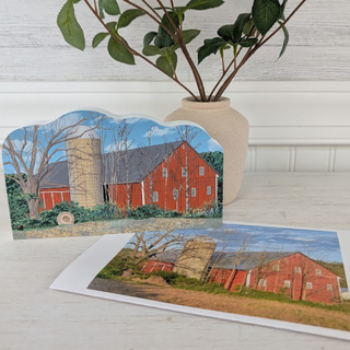A decorative cutout and matching print of a red barn scene with trees and a silo, styled on a white surface with a vase of greenery in the background.