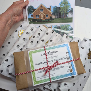 A hand holds a printed card above a wrapped package tied with red-and-white string. The package sits on white tissue paper printed with black paw prints, and the front label reads 'Cats Meow Village Homeowner's Association' with a name field.