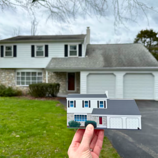 A miniature paper cutout model of a two-story house with a gray roof and white garage doors is held up by a person's hand in front of a real house on a lawn.