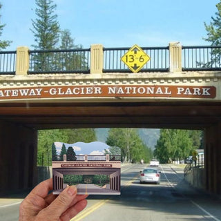 A hand holds a small wooden souvenir depicting the Gateway-Glacier National Park archway, positioned in front of a real outdoor scene of a road underpass with the same park signage visible above.