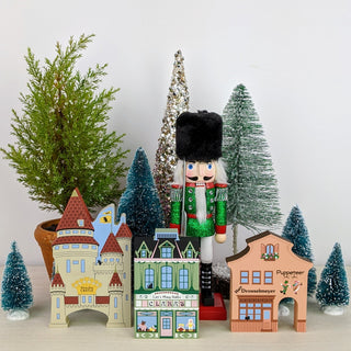 A festive holiday nutcracker displayed among miniature Christmas village buildings and small tree decorations against a plain light background.
