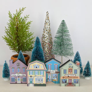 A set of decorative miniature Christmas village buildings and trees, including colorful house-shaped trees, small flocked pine trees, and a glittery silver tree, arranged on a light surface against a plain light background.