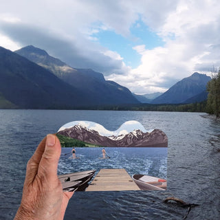 A hand holds a wooden replca of a mountain lake scene with a dock and two people stand-up paddleboarding in front of a real mountain landscape.