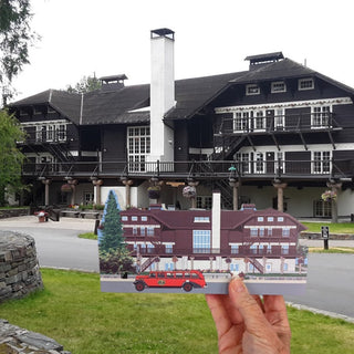 A hand holding a wooden replica of a large historic lodge building outdoors. The card shows a stylized red car and a white-and-brown building with a tall chimney.