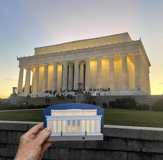 A hand holds a small wooden replica of the Lincoln Memorial in the foreground, with the real Lincoln Memorial photographed behind it at sunset.