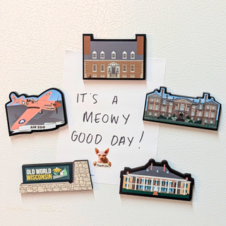 A set of illustrated refrigerator magnets arranged on a white textured surface around a handwritten note that reads 'IT'S A MEOW GOOD DAY!'. The magnets feature a brick building, an airplane at an air zoo, a classical building, a small dog portrait with 'Thank you' text, and a 'OLD WORLD WISCONSIN' sign.