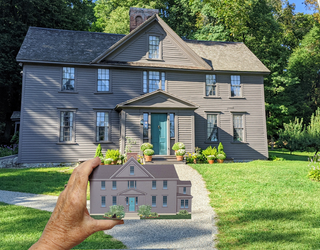 A hand holds a small wooden replica of a gray house with a teal front door in the foreground, while a large real house with matching gray siding and a teal door stands in the background on a green lawn with trees.