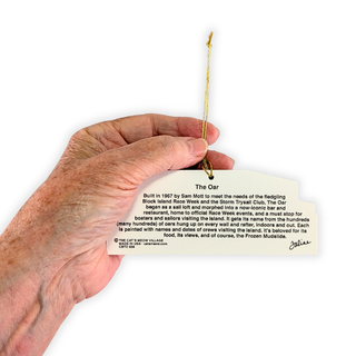 A hand holding a small printed tag or plaque with a gold hanging string. The tag reads 'The Oar' and includes descriptive text about a place or institution, photographed against a plain white background.