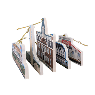 A set of four wooden Christmas ornaments shaped like historic buildings and storefronts, each printed with a different city or brand scene and hanging from a gold string on a white background.