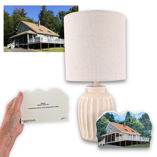 A cream-colored table lamp with a textured drum shade and ribbed ceramic base, shown alongside a hand holding a custom plaque, a small inset photo of a house, and a matching house-shaped plaque on a white background.