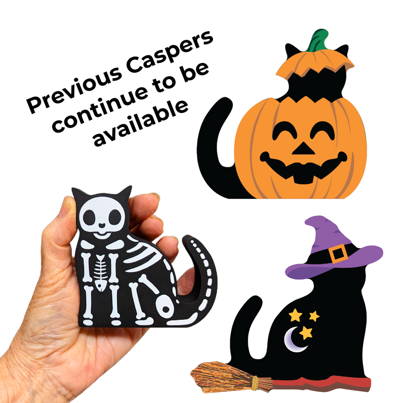A hand holds a small black-and-white skeleton cat Halloween ornament on a white background, with two larger pumpkin and witch cat cutout decorations shown above and the text 'Previous Caspers continue to be available.'.